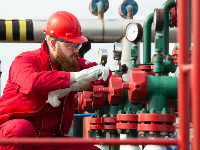 Engineer in red overalls, hard hat and safety glasses inspecting pressure gauges and valves during a UK commercial gas safety inspection
