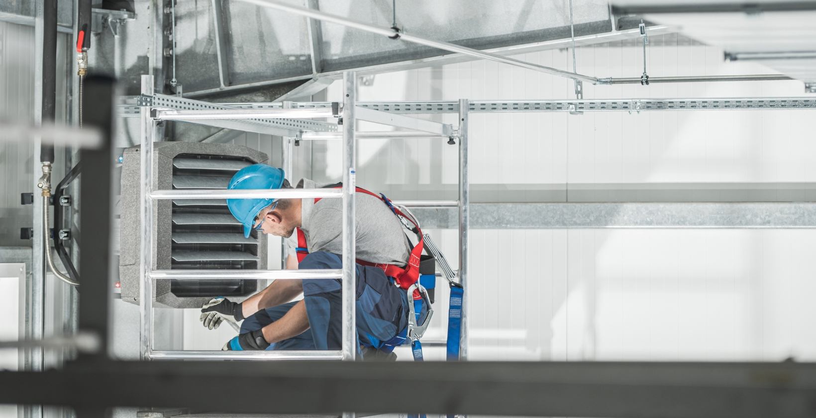Engineer in a hard hat and safety harness repairing a wall-mounted warehouse gas heater from an access tower in the UK